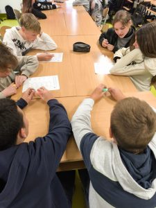 Grupa uczniów siedzi przy stole w klasie, wspólnie pracując nad kartkami z zadaniem. Uczniowie rozmawiają i piszą coś na arkuszach papieru, widać atmosferę współpracy.