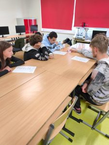 Grupa uczniów siedzi przy stole w sali komputerowej, wspólnie pracując nad kartkami z zadaniem. Na stole leżą arkusze papieru, uczniowie rozmawiają i analizują treść.