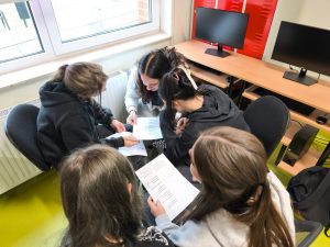Kilka uczennic siedzi w kręgu na krzesłach w sali komputerowej i wspólnie pracuje nad zadaniem zapisanym na kartkach. W tle widać monitory i czerwone szafki.
