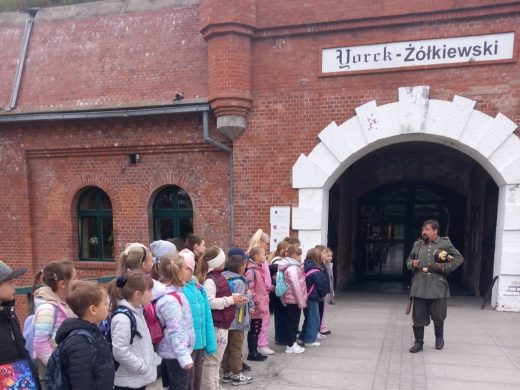 Grupa dzieci stoi w rzędzie przed ceglaną bramą z napisem „Yorck-Żółkiewski”. Przewodnik w historycznym stroju opowiada im o miejscu. Scena wygląda jak wycieczka edukacyjna do zabytkowego fortu.