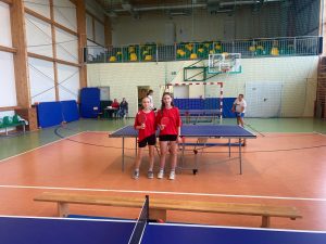 Na zdjęciu dwie dziewczyny w czerwonych koszulkach sportowych stoją przy stole do tenisa stołowego w sali gimnastycznej. Każda z nich trzyma rakietkę do ping-ponga. W tle widać sędziów siedzących przy stole oraz inną osobę przygotowującą się do gry.