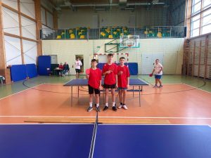 Na zdjęciu trzej chłopcy w czerwonych koszulkach sportowych stoją obok siebie przy stole do tenisa stołowego, każdy trzyma rakietkę. W tle widać sędziów przy stole oraz dwóch innych zawodników w białych koszulkach.
