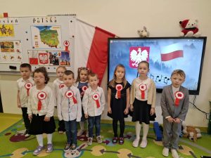 Grupa dzieci z oddziału przedszkolnego w odświętnych strojach stoi w sali lekcyjnej przed ekranem wyświetlającym flagę i godło Polski. Na tablicy obok znajdują się szkolne dekoracje związane z obchodami Święta Niepodległości.