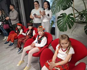 Kilka dzieci w ludowych strojach siedzi na czerwonych fotelach, a stojący obok dorośli biją brawo po występie podczas przeglądu pieśni patriotycznych.