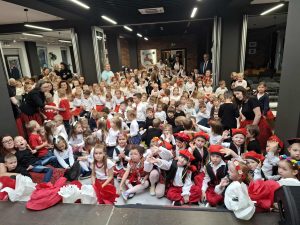 Sala wypełniona przedszkolakami w biało-czerwonych strojach oraz nauczycielami i rodzicami; dzieci siedzą przed sceną podczas szkolnego przeglądu pieśni patriotycznych.