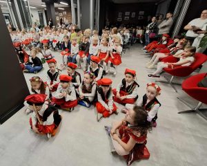 Dzieci z oddziału przedszkolnego w strojach ludowych siedzą z przodu sali, za nimi pozostali uczestnicy i publiczność oglądająca występy na przeglądzie pieśni patriotycznych.