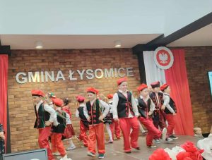 Dzieci w ludowych strojach prezentują układ taneczny na scenie udekorowanej biało-czerwonymi kwiatami; w tle widoczny napis „Gmina Łysomice” i symbol orła w barwach narodowych.