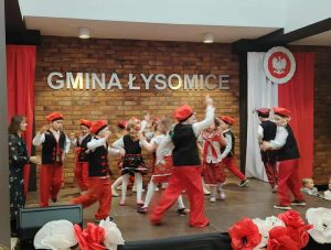 Grupa przedszkolaków w czerwonych spodniach, kamizelkach i czapkach oraz dziewczynki w ludowych spódniczkach wykonują taniec na scenie podczas przeglądu pieśni patriotycznych, obok nich stoi nauczycielka.