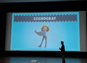 Na ekranie kinowym wyświetlona jest ilustracja postaci trzymającej plan filmowy, z napisem „Scenograf”. Na scenie stoi prowadzący zajęcia.