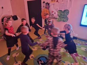 Dzieci w przebraniu na Halloween trzymają się za ręce i tańczą w kółku na dywanie w przedszkolnej sali. W tle widać dekoracje jesienne i halloweenowe.