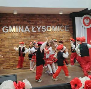 Grupa przedszkolaków w tradycyjnych biało-czerwonych strojach tańczy na scenie podczas występu w przeglądzie pieśni patriotycznych, na tle napisu „Gmina Łysomice” i dekoracji w barwach narodowych.