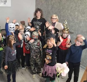 Grupa dzieci pozuje w halloweenowych przebraniach wraz z wychowawczynią i panią dyrektor w jej gabinecie. Dzieci mają stroje m.in. czarownic, mumii, wampirów i masek duchów, machają w stronę aparatu.