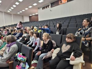 Grupa dzieci i opiekunów siedzi w dużej sali kinowej podczas edukacyjnego seansu o zawodach. Widać rzędy foteli i uczniów przygotowanych do zajęć.