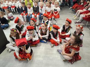 Grupa dzieci z oddziału przedszkolnego w biało-czerwonych strojach ludowych siedzi na podłodze i czeka na występ podczas szkolnego przeglądu pieśni patriotycznych.