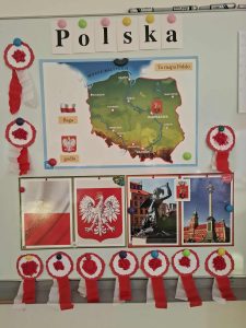 Tablica szkolna udekorowana materiałami związanymi z Polską, w tym mapą kraju, flagą, godłem oraz zdjęciami zabytków. Wokół tablicy zawieszone są biało-czerwone kotyliony wykonane przez dzieci.