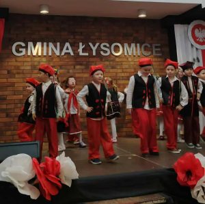 Dzieci w tradycyjnych ludowych strojach w biało-czerwonych barwach tańczą na scenie podczas przeglądu pieśni patriotycznych; w tle widoczny napis „Gmina Łysomice” oraz dekoracje w barwach narodowych.