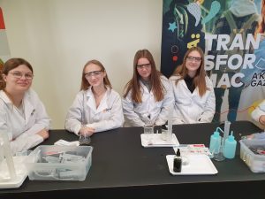 Cztery uczennice w fartuchach laboratoryjnych siedzą przy stole i przygotowuje się do wykonania doświadczenia chemicznego. Mają na sobie okulary ochronne. Na stole leżą cylindry miarowe, zlewki, butelki z wodą destylowaną i plastikowe pojemniki z przyborami. Za nimi widoczny jest baner edukacyjny.