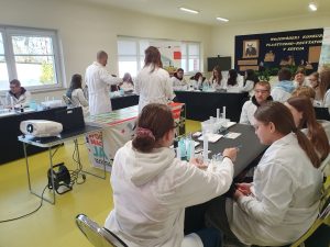 Uczniowie klasy ósmej w fartuchach laboratoryjnych siedzą przy stolikach w sali teatralnej i wykonują doświadczenia chemiczne. Na stołach znajdują się cylindry miarowe, probówki, pipety i zlewki. Nauczyciel w białym fartuchu prowadzi zajęcia. Z boku widoczny jest projektor, a w tle dekoracja dotycząca konkursu szkolnego.