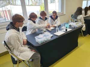 Kilku uczniów w fartuchach laboratoryjnych siedzi przy stole i pracuje wspólnie nad doświadczeniem chemicznym. Na stole znajdują się naczynia miarowe, zlewki, mały lejek oraz plastikowe butelki laboratoryjne. Za nimi widać duże okna i naturalne światło wpadające do sali.