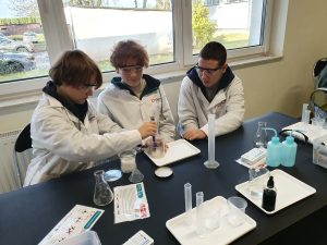 Troje uczniów w białych fartuchach wykonuje eksperyment chemiczny przy stole. Jeden uczeń odmierza substancję do naczynia laboratoryjnego, pozostali obserwują. Na stole leżą zlewki, kolby, cylindry miarowe oraz instrukcje do doświadczeń. W tle widać okno z widokiem na zieleń.