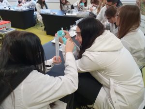 Grupa uczniów w białych fartuchach wykonuje doświadczenie chemiczne. Dwoje uczniów trzyma cylinder miarowy i plastikową butelkę z nakładką, koncentrując się na eksperymencie. Na stole widoczne są probówki, pipety i inne przyrządy laboratoryjne.