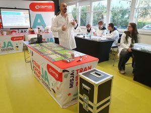 Prowadzący w fartuchu laboratoryjnym stoi przed uczniami i prowadzi zajęcia doświadczalne. Uczniowie siedzą przy stołach z materiałami laboratoryjnymi, a w tle widać ekran z prezentacją oraz kolorowe elementy dekoracyjne związane z tematyką naukową.