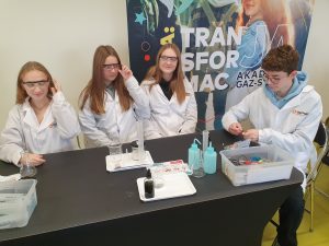 Czworo uczniów siedzi przy stole w fartuchach laboratoryjnych, a jedna osoba przygotowuje elementy eksperymentu. Uczniowie mają założone okulary ochronne, a na stole znajdują się cylindry miarowe, kolby, zlewki oraz zestaw przyborów w plastikowym pudełku. W tle widoczny baner akademii edukacyjnej.