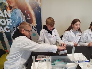 Troje uczniów w białych fartuchach laboratoryjnych siedzi przy stole i przygotowuje elementy potrzebne do eksperymentu chemicznego. Na stole znajdują się plastikowe pudełko z przyborami, cylindry miarowe oraz zlewki. W tle widoczny kolorowy baner wydarzenia edukacyjnego.