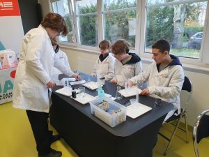 Uczniowie w fartuchach laboratoryjnych wykonują doświadczenie chemiczne przy stole. Jeden uczeń stoi i odmierza substancję do naczynia, pozostali siedzą i przygotowują sprzęt. Na stole widoczne są zlewki, pipety, cylindry miarowe oraz czarne buteleczki z odczynnikami, a w tle okna z widokiem na drzewa.