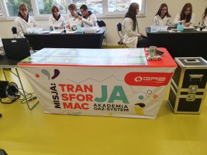 Stół demonstracyjny z banerem edukacyjnym „Misja: Transformacja. Akademia GAZ-SYSTEM”. Na stole znajduje się model pojazdu i przyrządy doświadczalne. W tle uczniowie w fartuchach laboratoryjnych pracują przy swoich stanowiskach, wykonując eksperymenty chemiczne.