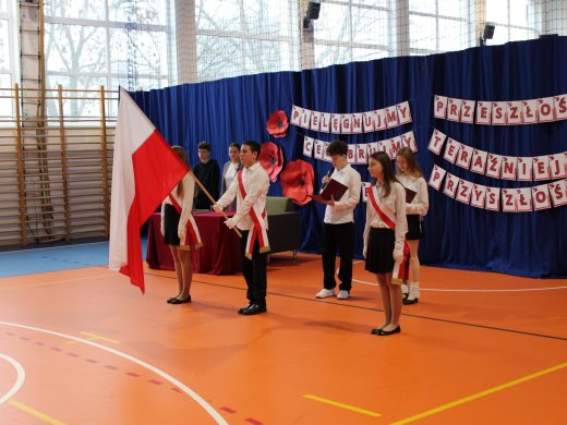 Poczet flagowy stoi podczas apelu zorganizowanego z okazji Święta Niepodległości na sali gimnastycznej. Uczeń stojący po środku trzyma przed sobą opuszczoną w dół flagę. W tle znajduje się dekoracja z biało-czerwonym napisem oraz dużymi papierowymi makami.