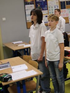 Uczniowie stoją przy swoich ławkach w klasie i biorą udział w odśpiewaniu hymnu narodowego z okazji Święta Niepodległości.