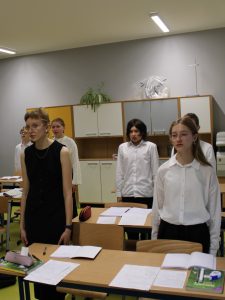 Uczniowie stoją przy swoich ławkach w klasie i biorą udział w odśpiewaniu hymnu narodowego z okazji Święta Niepodległości. Na ławkach widoczne są zeszyty i przybory szkolne.
