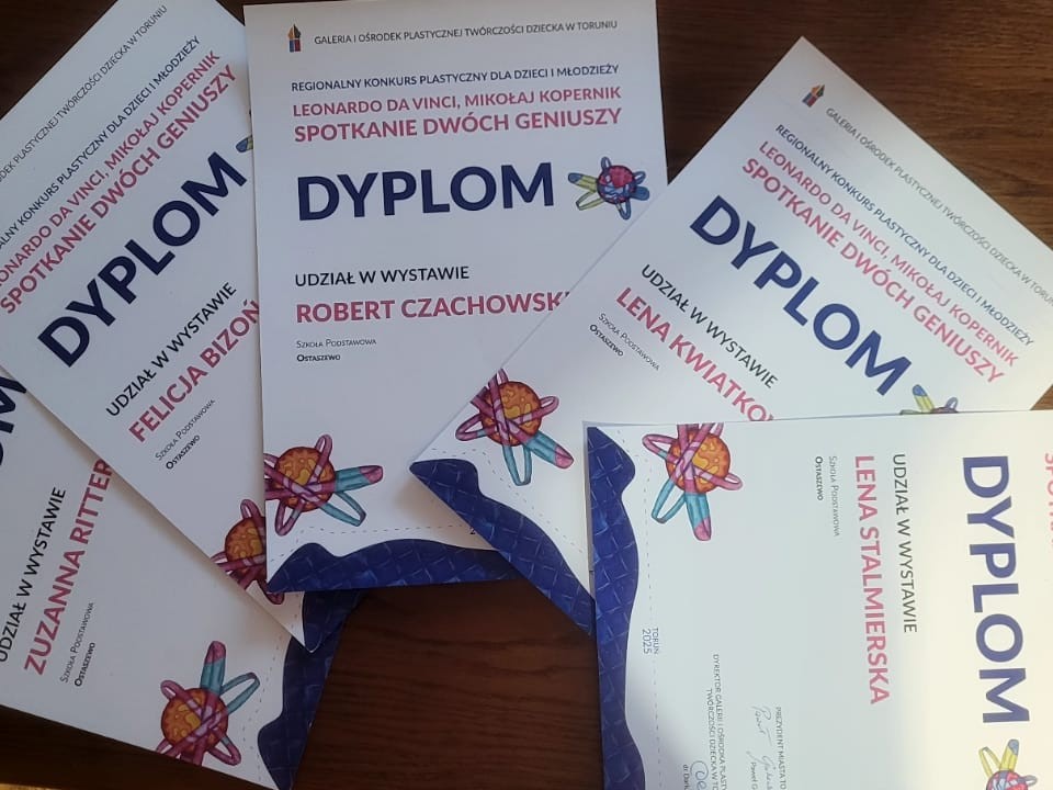 Na drewnianym blacie leży kilka dyplomów za udział w regionalnej wystawie plastycznej dla dzieci i młodzieży pt. ‘Spotkanie dwóch geniuszy’. Dyplomy częściowo na siebie zachodzą i zawierają imiona uczestników oraz kolorowe grafiki inspirowane motywami naukowymi.