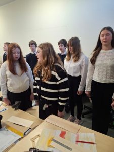Uczniowie stoją przy stołach w pracowni i uczestniczą w odśpiewaniu hymnu narodowego z okazji Święta Niepodległości. Na stołach leżą przybory szkolne i prace plastyczne.