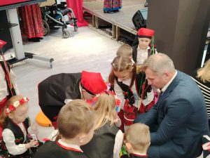 Dzieci w strojach ludowych siedzą i stoją w grupie przy scenie, rozmawiając z dorosłym mężczyzną w garniturze podczas przeglądu pieśni patriotycznych.