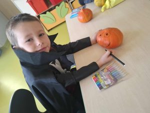Dziecko w halloweenowym kostiumie siedzi przy stoliku i rysuje uśmiechniętą buzię na małej pomarańczowej dyni za pomocą flamastra. Na stole leżą kolorowe pisaki.