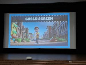 Na ekranie kinowym widoczna jest animowana postać stojąca na tle mieście z napisem „Green screen”. Obraz przedstawia technikę tworzenia cyfrowego tła w filmach.