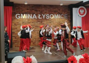 Przedszkolaki w kolorowych ludowych strojach tańczą na scenie podczas przeglądu pieśni patriotycznych; z boku stoi nauczycielka, a tło stanowi napis „Gmina Łysomice” oraz biało-czerwone dekoracje.