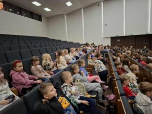 Uczniowie klas 0–3 siedzą w kinowej sali podczas edukacyjnej lekcji o zawodach filmowych. Dzieci oglądają prezentację na dużym ekranie, zajmując kilka rzędów foteli.