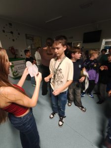 Uczniowie rozmawiają i biorą udział w zabawie andrzejkowej na korytarzu szkolnym. Na pierwszym planie widoczna jest uczennica trzymająca przed sobą wycięte z papieru serce. Na przeciwko niej stoi uczeń patrzący w jego stronę.