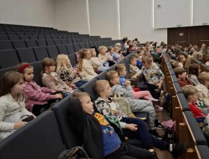 Dzieci z klas 0–3 siedzą w kinowej sali na lekcji o zawodach. Uczniowie zajmują rzędy foteli, patrząc w stronę ekranu.