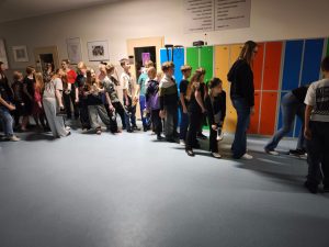 Grupa uczniów ustawia buty w rzędzie na szkolnym korytarzu podczas tradycyjnej wróżby andrzejkowej. W tle widać kolorowe szafki stojące na korytarzu.