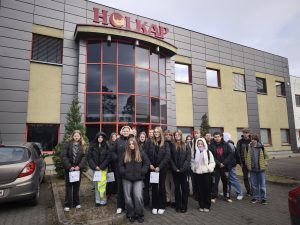 Grupa uczniów klasy VIII stoi przed budynkiem firmy HOLKAP należącej do Bydgoskiego Klastra Przemysłowego; w tle widoczna jest nowoczesna fasada budynku z czerwonymi ramami okien.