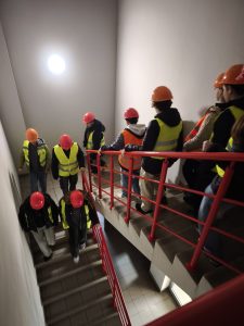 Grupa uczniów w kaskach ochronnych i odblaskowych kamizelkach schodzi po klatce schodowej o czerwonych poręczach podczas zwiedzania zakładu produkcyjnego firmy HOLKAP.