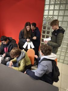 Uczniowie klasy VIII siedzą i stoją w sali firmy HOLKAP; trzymają w rękach torby upominkowe i notatki, a w tle widoczna jest czerwona ściana oraz szklana ściana z pustaków.