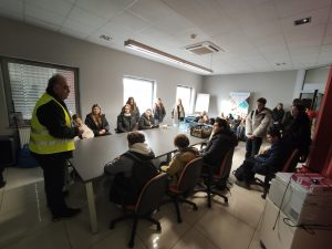 Grupa uczniów i uczennic klasy VIII uczestniczy w spotkaniu informacyjnym w sali firmy HOLKAP; pracownik w kamizelce odblaskowej omawia specyfikę pracy w branży narzędziowej.