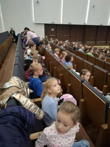 Uczniowie klas 0–3 siedzą w kinie, czekając na rozpoczęcie lekcji o zawodach. Dzieci rozmawiają między sobą i patrzą w stronę ekranu.