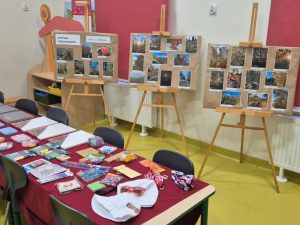 Trzy tablice na sztalugach prezentujące zdjęcia przyrodnicze zgłoszone do szkolnego konkursu fotograficznego. Na stole przed nimi rozłożone są nagrody i upominki dla uczestników, takie jak artykuły plastyczne i drobne gadżety.