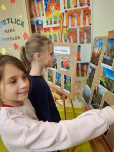 Dwie dziewczynki stoją przy sztaludze z fotografiami konkursowymi i wskazują jedno ze zdjęć. W tle widać barwne prace plastyczne przedstawiające jesienne drzewa.